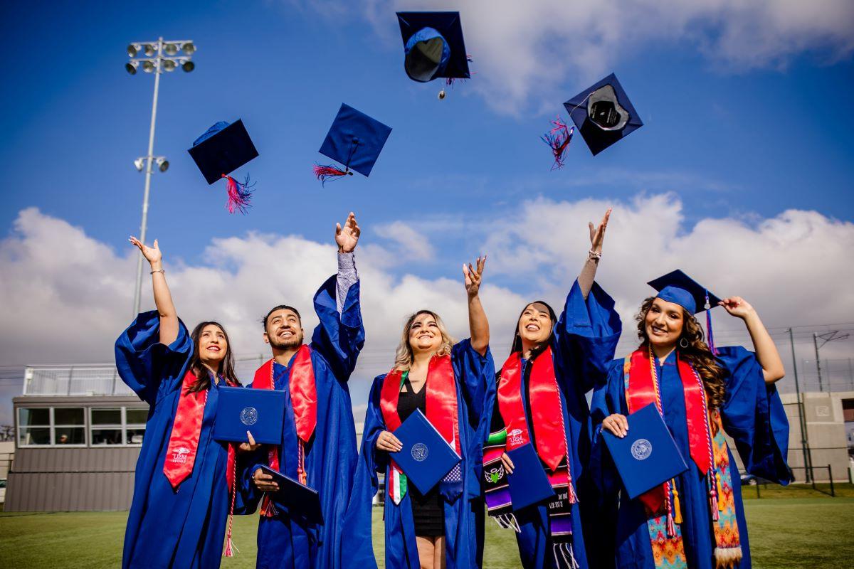 Five graduates in regalia throwing their caps in the air.